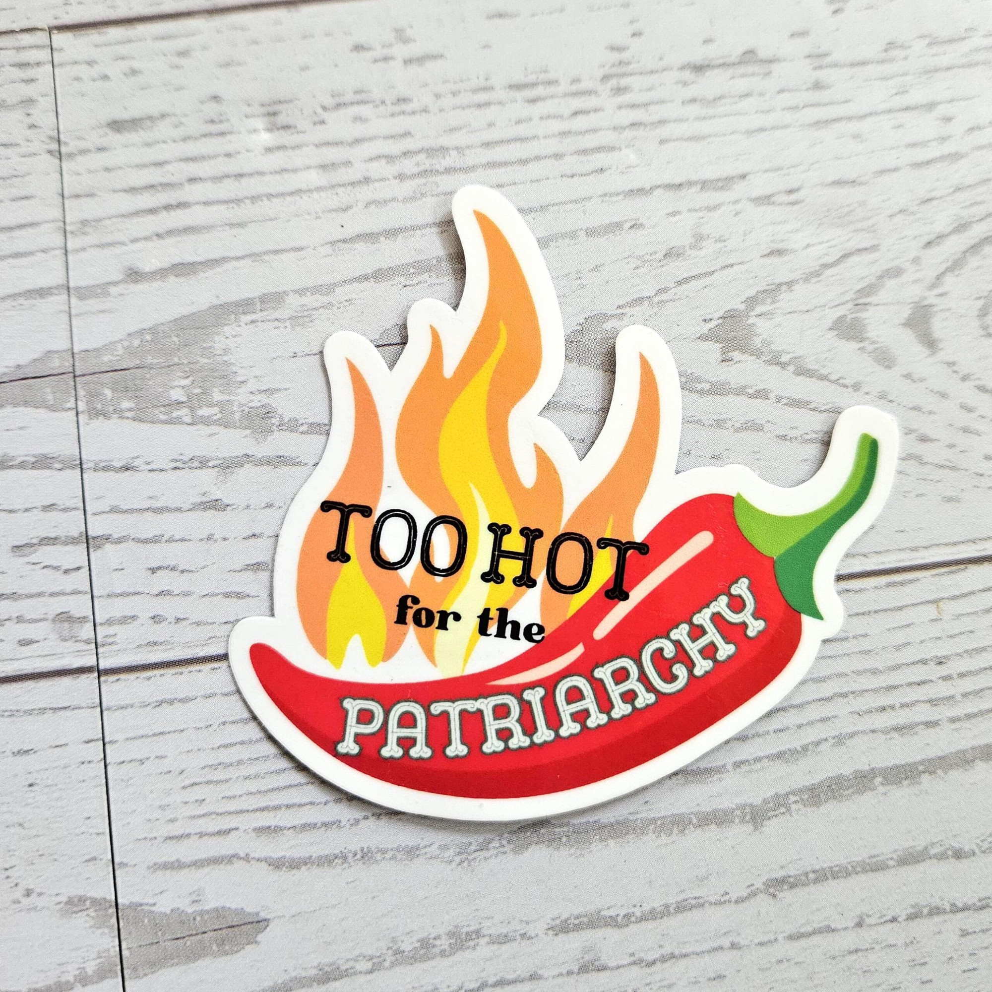 Too Hot for the Patriarchy Sticker. a sticker of a chili pepper with flames on a wood surface